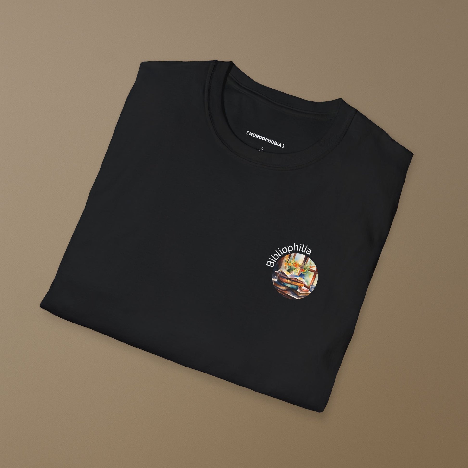 Folded black T-shirt with a graphic design of a watercolor painting with pile of books and flowers near the window and text "Bibliophilia"  on a brown background