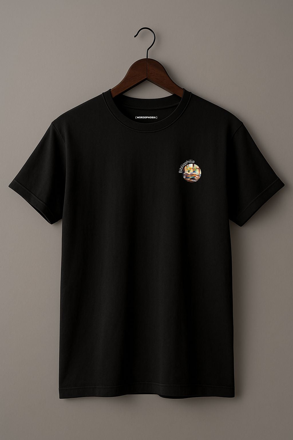 Black t-shirt with a graphic design of a watercolor painting with pile of books and flowers near the window and text "Bibliophilia" on a wooden hanger against a plain background