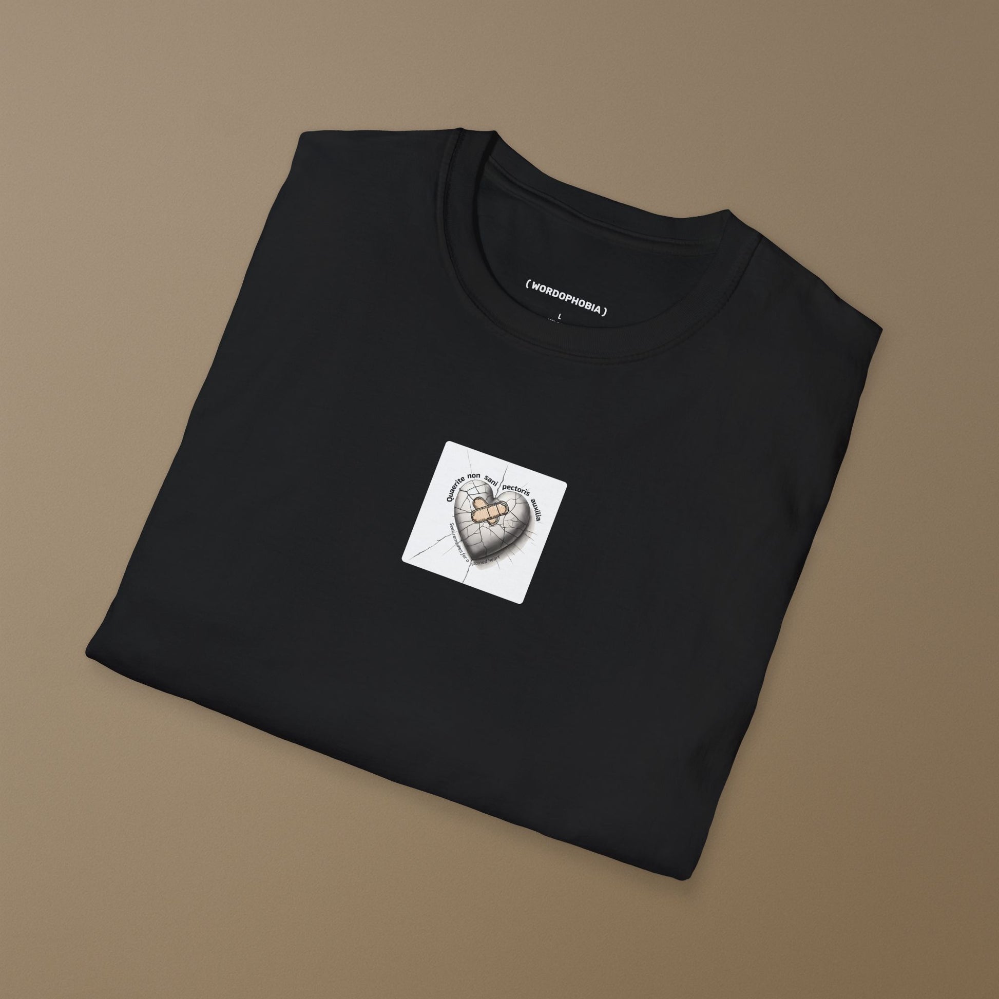 Folded black T-shirt with a small white patch featuring a design of heart with a bandage and cracked surface, featuring the Latin phrase “Quaerite non sani pectoris auxilia,” meaning “Seek remedies for a sick heart,” on a brown background