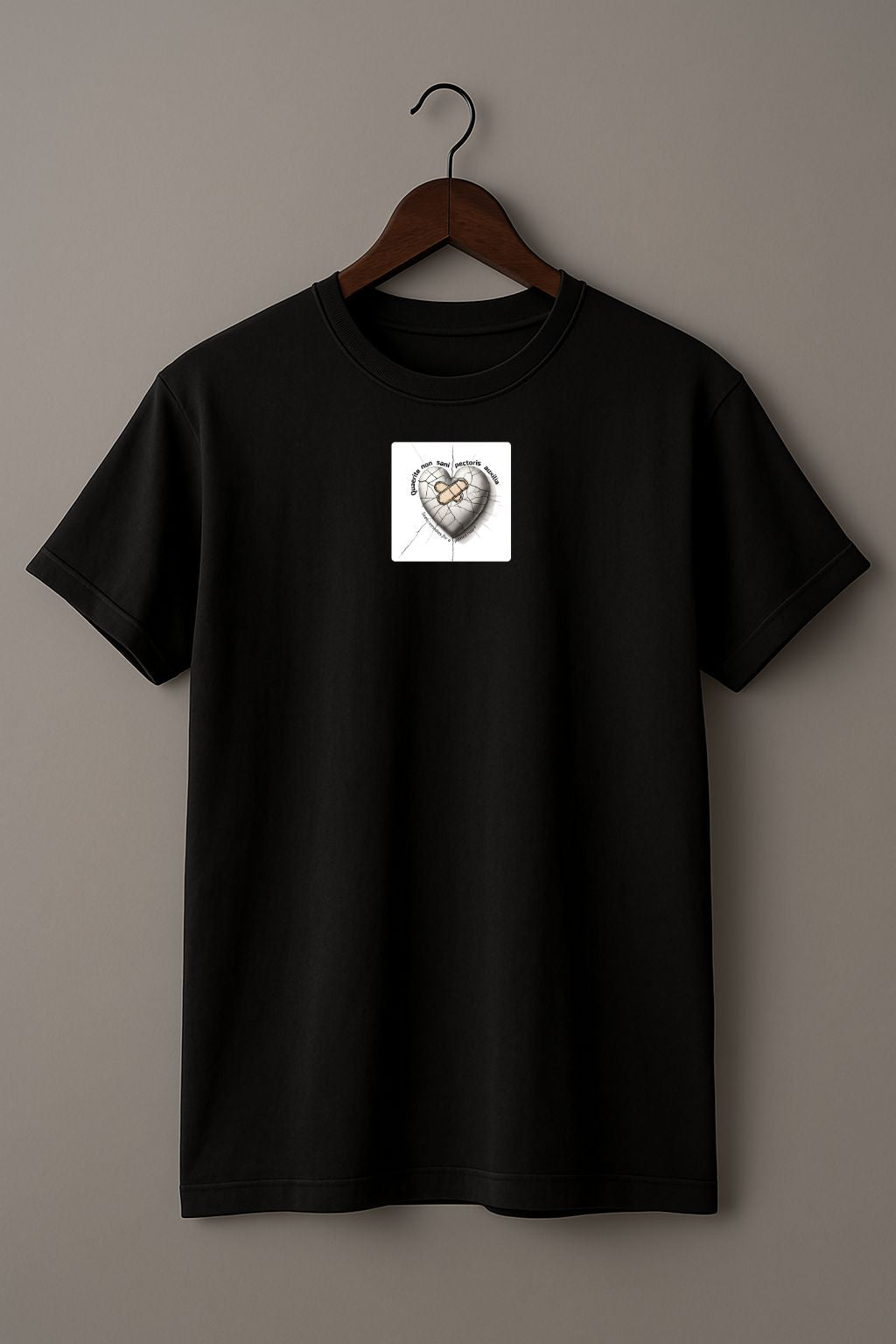 Black T-shirt with a small white patch featuring a design of heart with a bandage and cracked surface, featuring the Latin phrase “Quaerite non sani pectoris auxilia,” meaning “Seek remedies for a sick heart,” on a wood hanger against a plain background