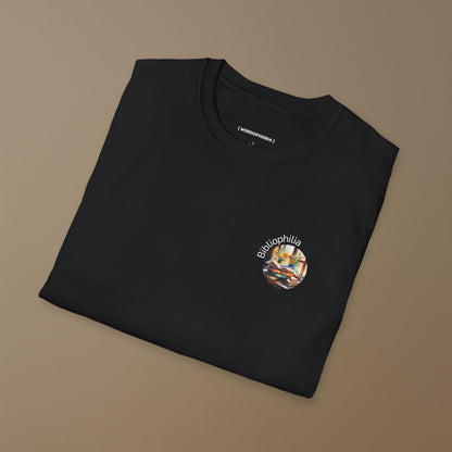 Folded black T-shirt with a graphic design of a watercolor painting with pile of books and flowers near the window and text "Bibliophilia"  on a brown background