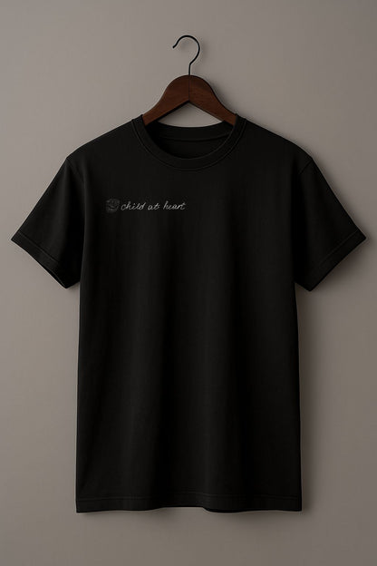 Black t-shirt with hand drawn kids picture of the funny face and the hand written text "child at heart" on a wooden hanger against a plain background