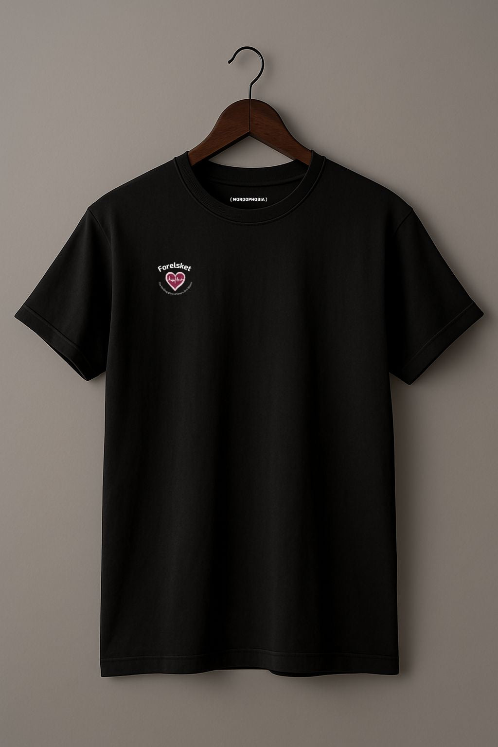 Black t-shirt with a small design of a heart with a beat and texts  "Forelsket" and "The thrilling glow of love's first bloom" on a wooden hanger against a plain background