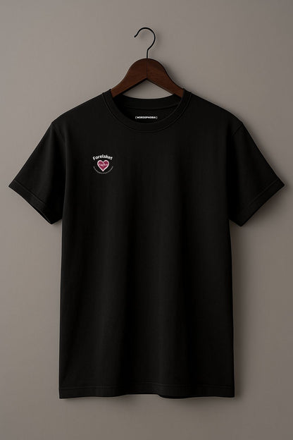 Black t-shirt with a small design of a heart with a beat and texts  "Forelsket" and "The thrilling glow of love's first bloom" on a wooden hanger against a plain background