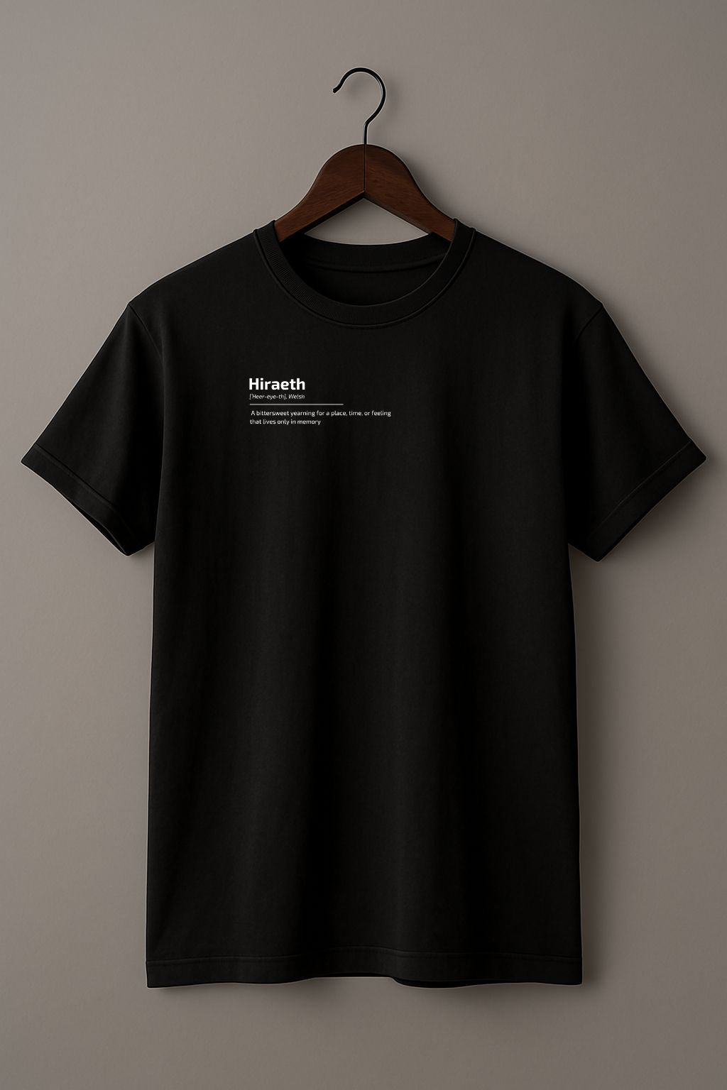 Black t-shirt with graphic design of the word HIRAETH ([ˈHeer-eye-th], Welsh) and its definition "A bittersweet yearning for a place, time, or feeling that lives only in memory" on a wooden hanger against a plain background