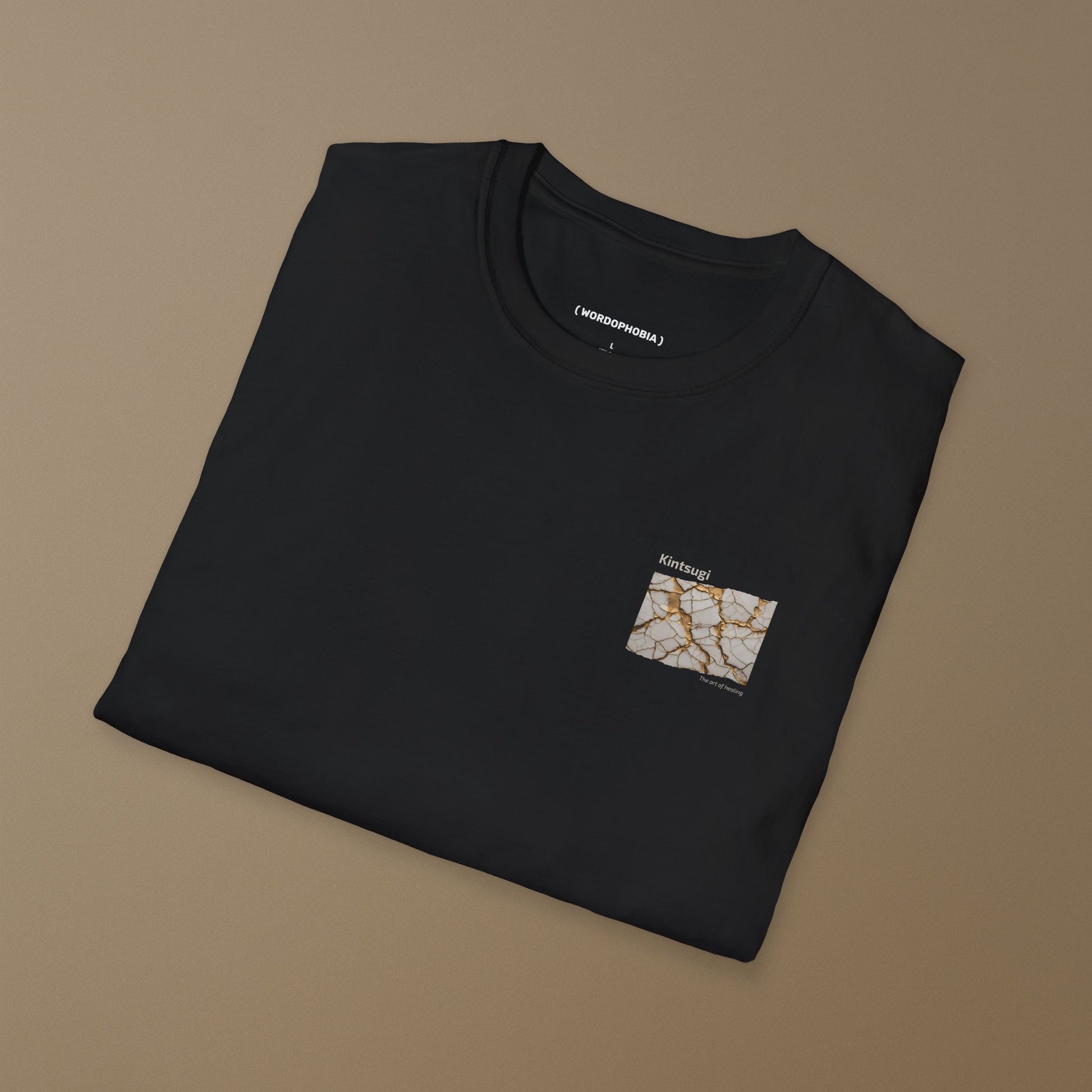 Folded black t-shirt with a small graphic of Kintsugi art with gold repair on a cracked white surface, text 'Kintsugi The art of healing' above on a brown background