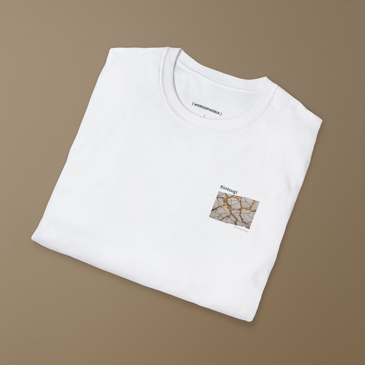 Folded white t-shirt with a small graphic of Kintsugi art with gold repair on a cracked white surface, text 'Kintsugi The art of healing' above on a brown background