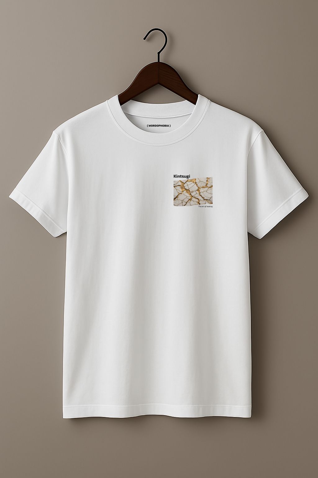 White t-shirt with a small graphic of Kintsugi art with gold repair on a cracked white surface, text 'Kintsugi The art of healing' above on a hanger against a beige background