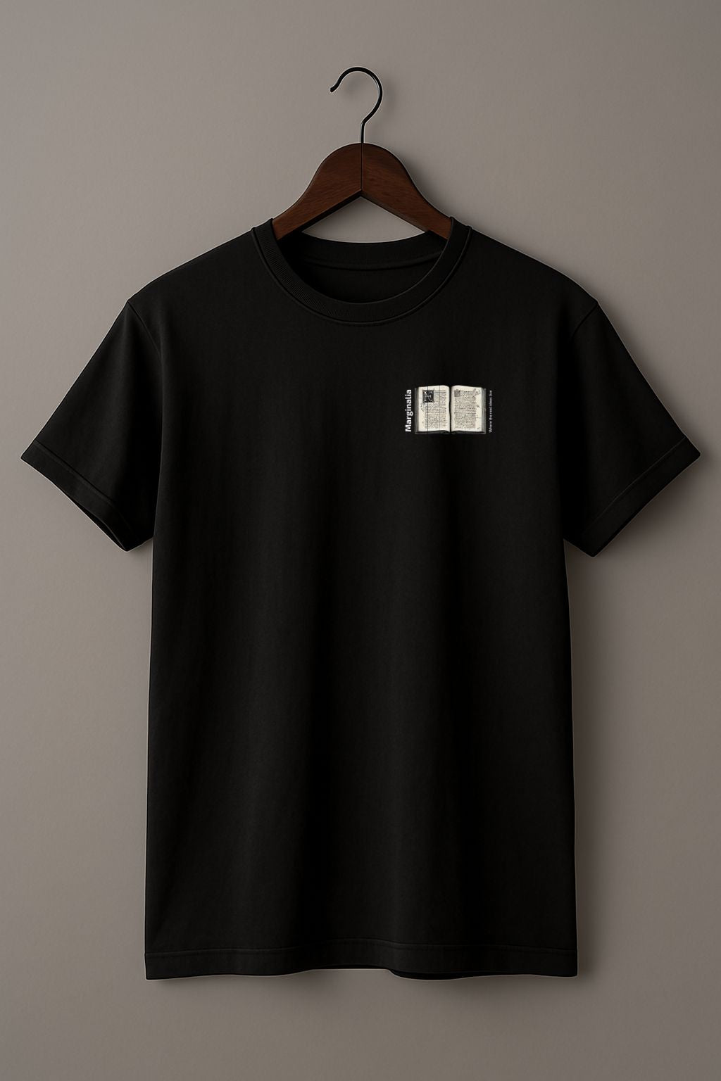 Black t-shirt with a small graphic of Enlarged design of open old book with text "Marginalia" and "Where the real ideas live" on a hanger against a beige background