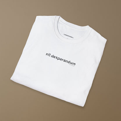Folded white T-shirt with the Latin phrase “Nil Desperandum,” meaning “Never Despair,” printed in cracked typeface on the beige background