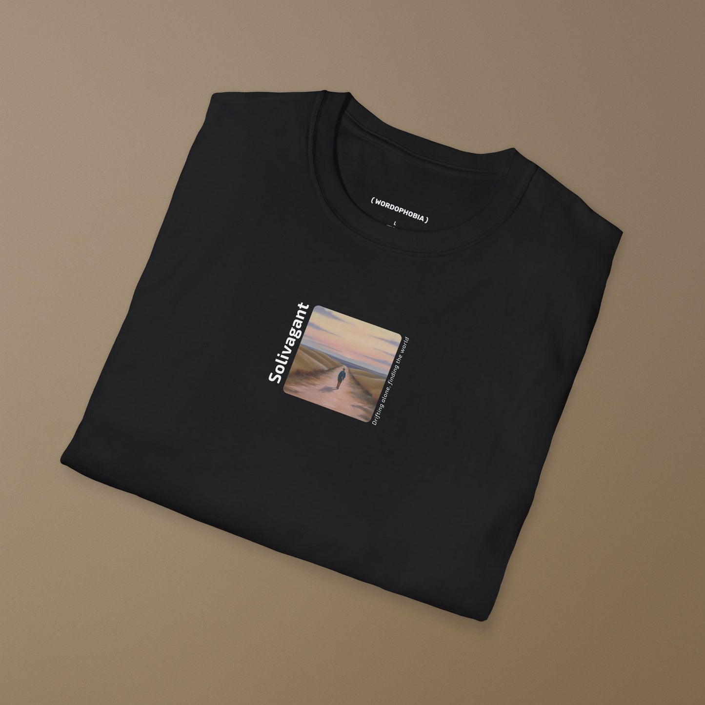 Folded black t-shirt with the word “Solivagant,” text “Drifting alone, finding the world,” and watercolor art of a lone traveler walking between hills on a beige background