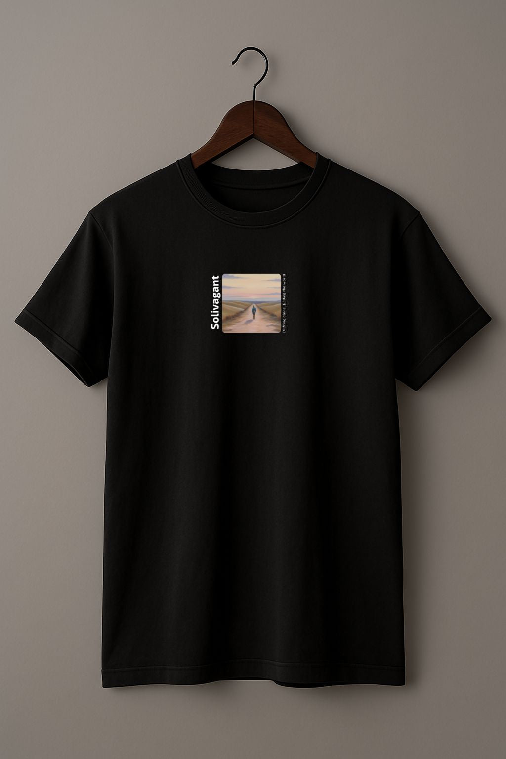 Black T-shirt with the word “Solivagant,” text “Drifting alone, finding the world,” and watercolor art of a lone traveler walking between hills on a wood hanger on a beige background