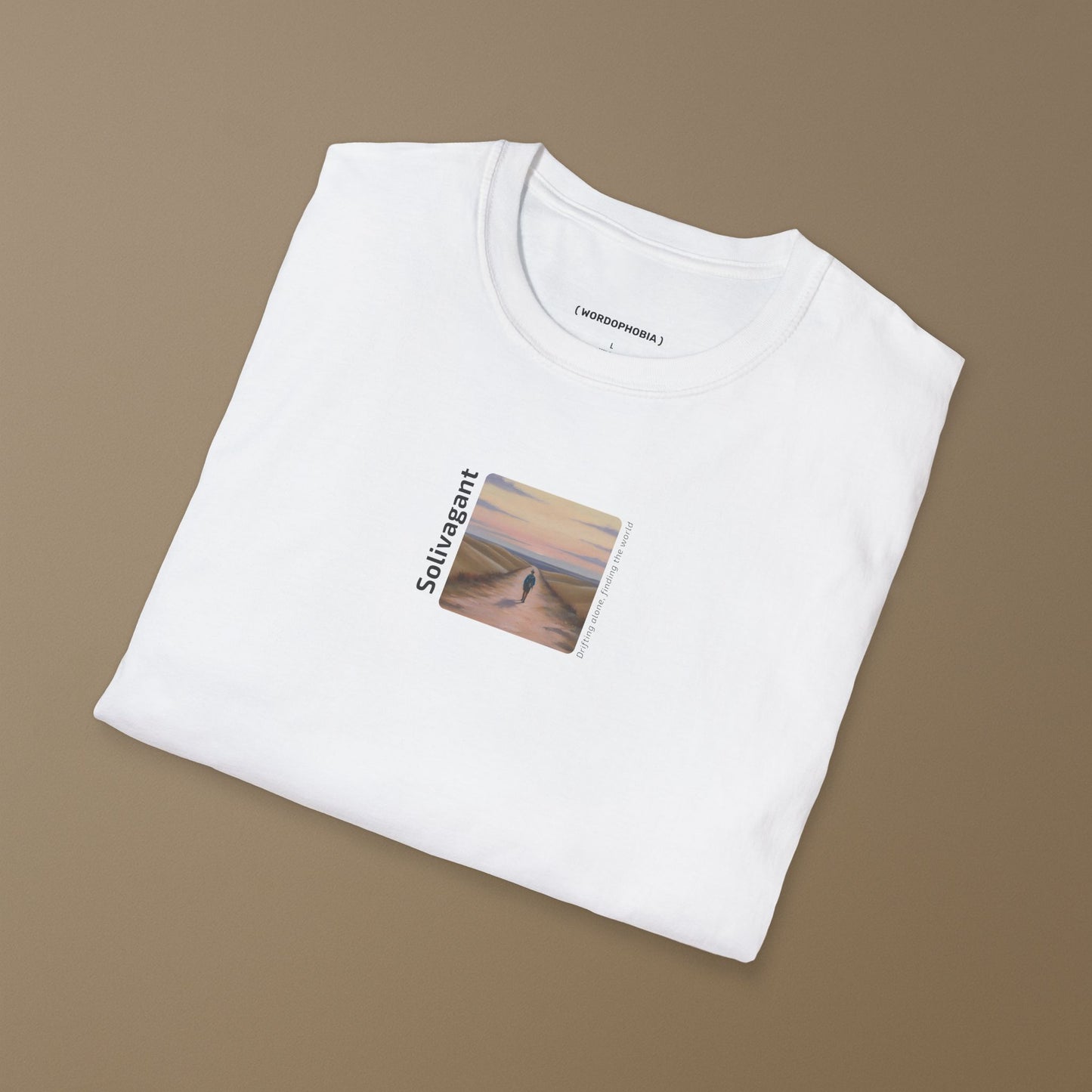 Folded white t-shirt with the word “Solivagant,” text “Drifting alone, finding the world,” and watercolor art of a lone traveler walking between hills on a beige background