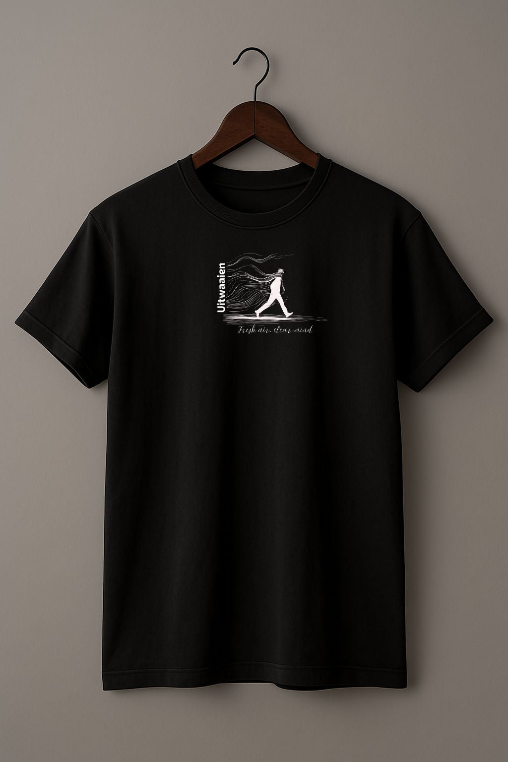 Black t-shirt featuring the Dutch word “Uitwaaien,” meaning “fresh air, clear mind,” with a small hand-drawn figure walking into the wind on the wood hanger on a beige background
