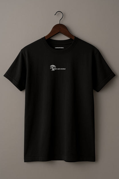 Black t-shirt with a small white design of an icon with 'Wabi-sabi mindset' text on a wooden hanger against a plain background